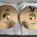 Dva mosazné činely typu hi-hat položené na bílé podložce. Horní činel má nápis TOP HI HAT a logo Zildjian. Spodní činel má nápis BOTTOM HI HAT a výra…