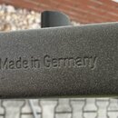 Detail šedé kovové trubky s matným povrchem. Na povrchu je vyražen nápis Made in Germany. V levé dolní části jsou vidět drobné známky opotřebení v po…