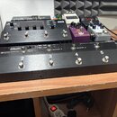 Na fotografii je několik kytarových efektů a spínacích jednotek na dřevěném stole. V popředí je černý kovový spínací pedalboard s šesti stisknutelným…
