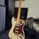 Bílá elektrická kytara typu Stratocaster s červeným želvovinovým pickguardem a javorovým hmatníkem. Na hlavě je viditelné logo Fender. Nástroj je umí…