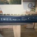 Černý digitální klavír s bílými a černými klávesami. Na zadní straně je bílý nápis KURZWEIL PC3 LE 6. V pravé části jsou umístěny konektory a ovládac…