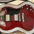 Červená elektrická kytara v tvaru SG položená v otevřeném kufru s bílou vnitřní výstýlkou. Nástroj má dva humbuckery, černý pickguard a čtyři ovládac…