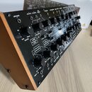 Černý analogový syntetizátor v dřevěném šasi. Na předním panelu jsou vidět různé ovládací knofíky, zapuštěné konektory a nápisy jako LFO RATE, LFO OU…