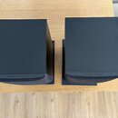 Dva černé, kostkovité reproduktory umístěné na dřevěném stole. Na spodní hraně obou přístrojů je nápis Bowers & Wilkins. Povrch je matný a v dobrém s…