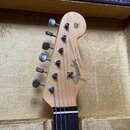 Hlava elektrické kytary z přírodního dřeva s černým hрифem. Na hlavě je černé logo Fender a nápis STRATOCASTER, pod kterým jsou menší texty: „WITH SY…