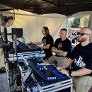 Fotografie zachycuje tři muže u pultů s audio technikou pod stříškou stanu. V popředí je digitální mixovací pult s modře podsvíceným displejem a posu…