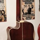 Zadní strana akustické kytary v tmavě červené barvě s bílým lemováním hran. Kytara má vyřezaný přístup k tělu (cutaway) a je umístěna na nástěnním dr…