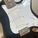 Černá elektrická kytara typu Stratocaster s bílým pickguardem a hnedým hmatníkem. Nástroj má tři bílé snímače s jednoduchou cívkou a ovládací knoflík…