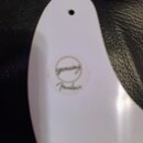 Bílá plastová pickguard s dvěma viditelnými otvory pro šrouby. Uprostřed je nalepený kulatý štítek s nápisy „genuine“ a „Fender“. Povrch je hladký a…