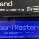 Černý elektronický modul s bílými nápisy Roland, DRUM SOUND MODULE TD-30 a SuperNATURAL. Modrý displej zobrazuje text DRUM KIT, Pear l Masters a v do…