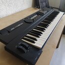 Černý digitální syntezátor s klávesnicí, umístěný na dřevěném stole. Na levé straně je bílý nápis YAMAHA YS200 a pod ním menší text DIGITAL SYNTHESIZ…