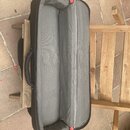 Otevřený černý polstrovaný pouzdro na hudební nástroj s šedým vnitřním povrchem. Pouzdro má na boku černé madlo a červené detaily u zipu. Na fotograf…