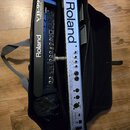 V černé přepravní tašce jsou umístěny dvě zařízení značky Roland. Vlevo je černý přístroj s nápisy Roland a VB-99 V-Bass System. Vpravo je stříbrný p…