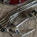 Fotografie zobrazuje chromovaný kovový hardware, konkrétně bubenový stojan a sadu pěti kovových tyčí s černými koncovkami. Některé z tyčí i rukojeť s…