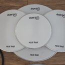 Pět bílých kruhových bubnových padů značky drum-tec. Každý pad má nápisy drum-tec v horní části a real feel ve spodní části. Povrchy padů jsou hladké…