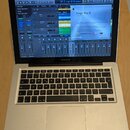 Stříbrný notebook MacBook Pro s otevřeným displejem. Na obrazovce je spuštěn program Logic Pro X verze 10.4.6 s otevřeným projektem a několika okny p…