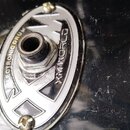 Detailní pohled na kovový oválný panel s konektorem, který je přišroubován k černé, poškrábané ploše. Na panelu je vyražen nápis ELECTRONIC DRUM a zn…