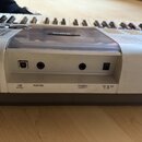 Detail zadní části bílé klávesové nástroje. Jsou vidět čtyři konektory s popisky USB, SUSTAIN, PHONES/ OUTPUT a DC IN 12V. Na horní části je viditeln…