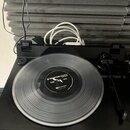 Černý gramofon s přeloženou stahovací raménkem. Na talíři je položena průhledná vinylová deska s černým středovým štítkem, na kterém je bílý nápis TR…