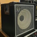 Černý basový reproduktor v bedně potažené černým materiálem s plastickými ochrániči rohů. Přední strana je kryta stříbrnou kovovou mřížkou, v jejím h…