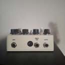 Bílá kytarový efekt s čtyřmi černými otočnými knoflíky. Na přední straně jsou tři konektory s popisky INPUT, REMOTE GAIN a OUTPUT. Zařízení vykazuje…