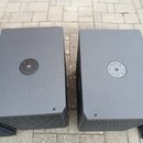 Dvě černé, hranaté reproduktorové bedny s texturovaným povrchem. Každá bedna má v horní části kruhový kovový prvek s šesti šrouby a na přední straně…