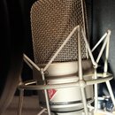 Kondenzátorový mikrofon s kovovou síťovkou a šedým tělem, umístěný v odpružívácím držáku (shock mount). Na těle je vidět červené kosočtvrý logo s náp…