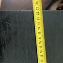 Detailní záběr na černý povrch s viditelnou strukturou dřeva. Vedle něj je přiložen žlutývstupní metr s černými a červenými číslicemi od 10 do 26. Po…