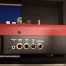 Detail zadní strany červeného hudebního přístroje s černým spodním panelem. Jsou zde vidět konektory s popisky MONITOR IN, CONTROL PEDAL, RIGHT OUT,…