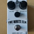 Bílý efekt pedál s nápisem THE WHITE ELK a pod ním SMALL FOOT. Na horní části jsou tři černé oválkové knoflíky s popisky VOLUME, TONE a DISTORTION. V…