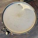 Fotografie zachycují pohled shora na buben s bílou membránou. Na horní části membrány je černý nápis REMO Ambassador EMPEROR BATTER MADE IN U.S.A. Po…