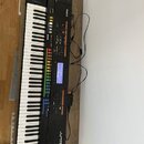 Černý digitální syntezátor s klaviaturou, umístěný na dřevěném stole. Na přístroji jsou viditelná loga a nápisy Roland a JUPITER-80. Ovládací panel o…