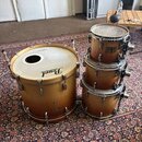 Sada bubnů v hnědém dřevěném provedení. Velký basový buben má na bílém blaně logo Pearl a v horní části vyřízný otvor. Vedle něj jsou tři stohované t…