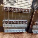 Detailní pohled na mechanismus s kovovými vřeteny a strunami, pravděpodobně součásti steel guitaru. Viditelný je kovový most a dvě řady hnědých vřete…