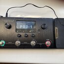 Černý efektový procesor s kovovým povrchem a nápisem AMPERO Amp Modeler / Effects Processor. Na displeji je zobrazen text P03-1 a Sick Lead, vedle js…