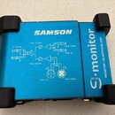 Modrý kovový box s černými gumovými nožičkami. Na horní straně je bílý nápis SAMSON a svisle orientovaný text G-monitor MONITOR/HEADPHONE AMP. Uprost…