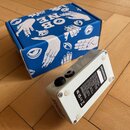 Bílá efektová pedál v kovovém šasi s dvěma vstupy/výstupy na straně. Na spodní straně je černá nálepka s technickým textem a symboly CE, RoHS. Vedle…