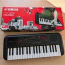 Na fotografii je černý digitální klavír Yamaha PSS-A50 a jeho původní červenobílá krabice. Na přístroji i na obalu je viditelné logo YAMAHA a označen…