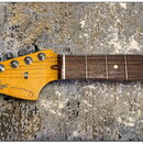 Fotografie zachycuje hmatník a hlavu elektrické kytary s přírodním dřevěným povrchem. Na hlavě je čitelné logo Fender a nápis JAZZMASTER, CORONA CALI…