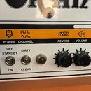 Detail ovládacího panelu kytarového zesilovače značky Orange. Panel je bílý s oranžovým pruhem, na kterém jsou černé nápisy POWER, CHANNEL, REVERB, V…