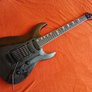 Černá elektrická kytara s tmavým hnedým hmatníkem a špičatou hlavou. Na hlavě je žlutý nápis KRAGLER. Nástroj má dva humbuckery, jeden jednosmykový s…