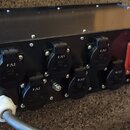 Černý kovový rozdělovač napájení s řadou kulatých konektorů. Na konektorech jsou viditelné nápisy F/L2, E/L1, C/L2, D/L3 a B/L1, přičemž u některých…