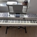 Stříbrný digitální klavír s černými a bílými klávesami, umístěný na černém X-stojanu. Na horním panelu je uprostřed logo YAMAHA a vpravo nápis Clavin…