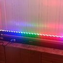 Černý lineární LED panel s řadou svítidel, která září v barvách modré, zelené a červené. Na spodní části panelu je digitální displej s červeným nápis…