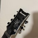 Černá hlava kytary s bílým logem Ibanez a nápisem Artist v kurzívě. Pod ním je menší svislý nápis SERIES. Na hlavě jsou vidět šest černých ladiček a…