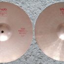Dva mosazné činely typu hi-hat položené vedle sebe na bílém koberci. Oba mají červené nápisy a logo Paiste 2002. Levý činel má nápis TOP MEDIUM HI-HA…