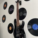 Černá elektrická kytara s tmavým hnedým hmatníkem, umístěná ve stěnovém držáku. Tělo je lesklé a vybaveno dvěma humbuckery, dvěma potenciometry a jed…