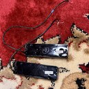 Dvě černé elektrické kytarové snímače s připojenými černými kabely. Na horním snímači je vyražené logo ve tvaru písmena A v kruhu a bílá nálepka s ná…