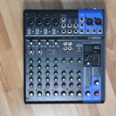 Černý mixovací pult s modrými prvky a bílými nápisy. Na přístroji je viditelné logo YAMAHA a označení modelu MG10XU MIXING CONSOLE. V pravé části se…