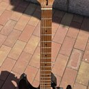 Elektrická kytara s tónem sunburst a bílým pickguardem. Na hlavě hředu je viditelný nápis Ibanez. Kytara má hnědý hmatník a je umístěna na černém sto…